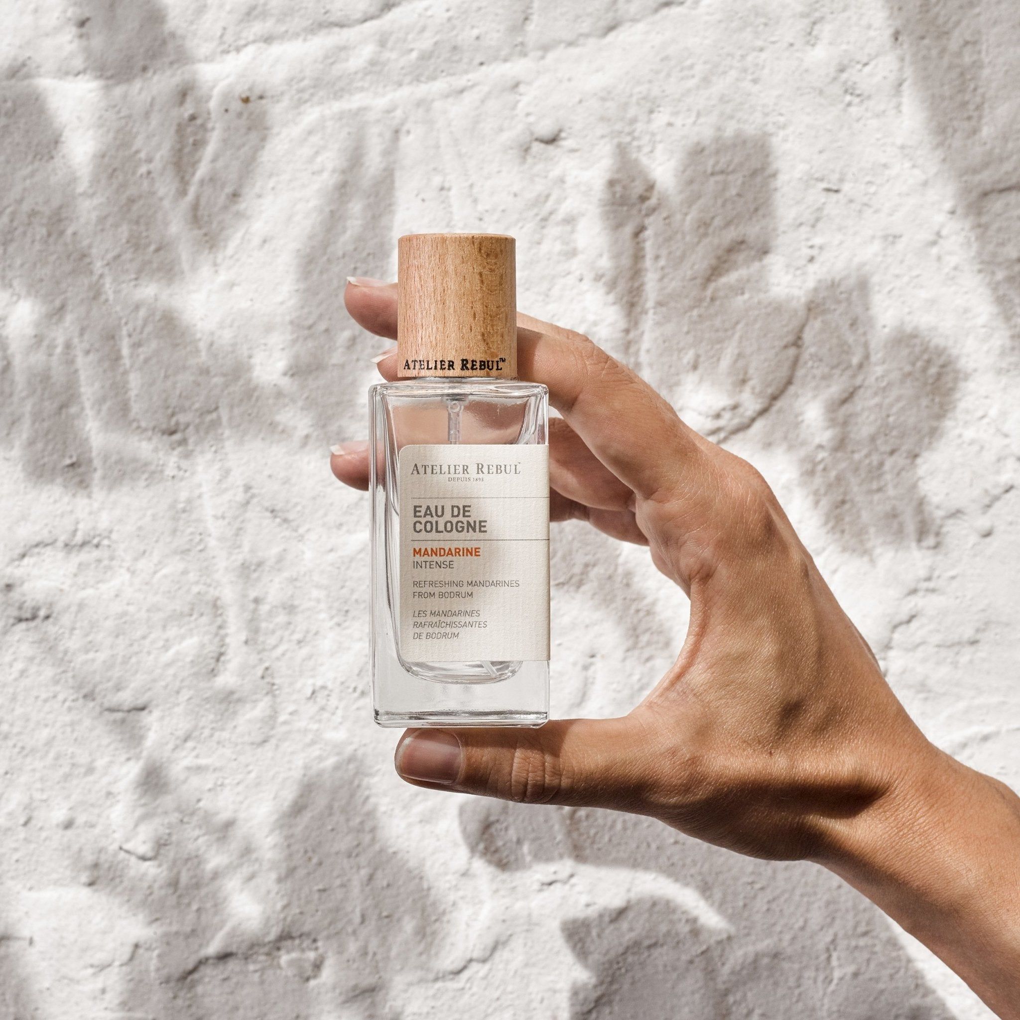 Klarer Glasflakon mit Holzverschluss. Etikett mit Produktinformationen. Hand hält Flakon vor weißer Wand.