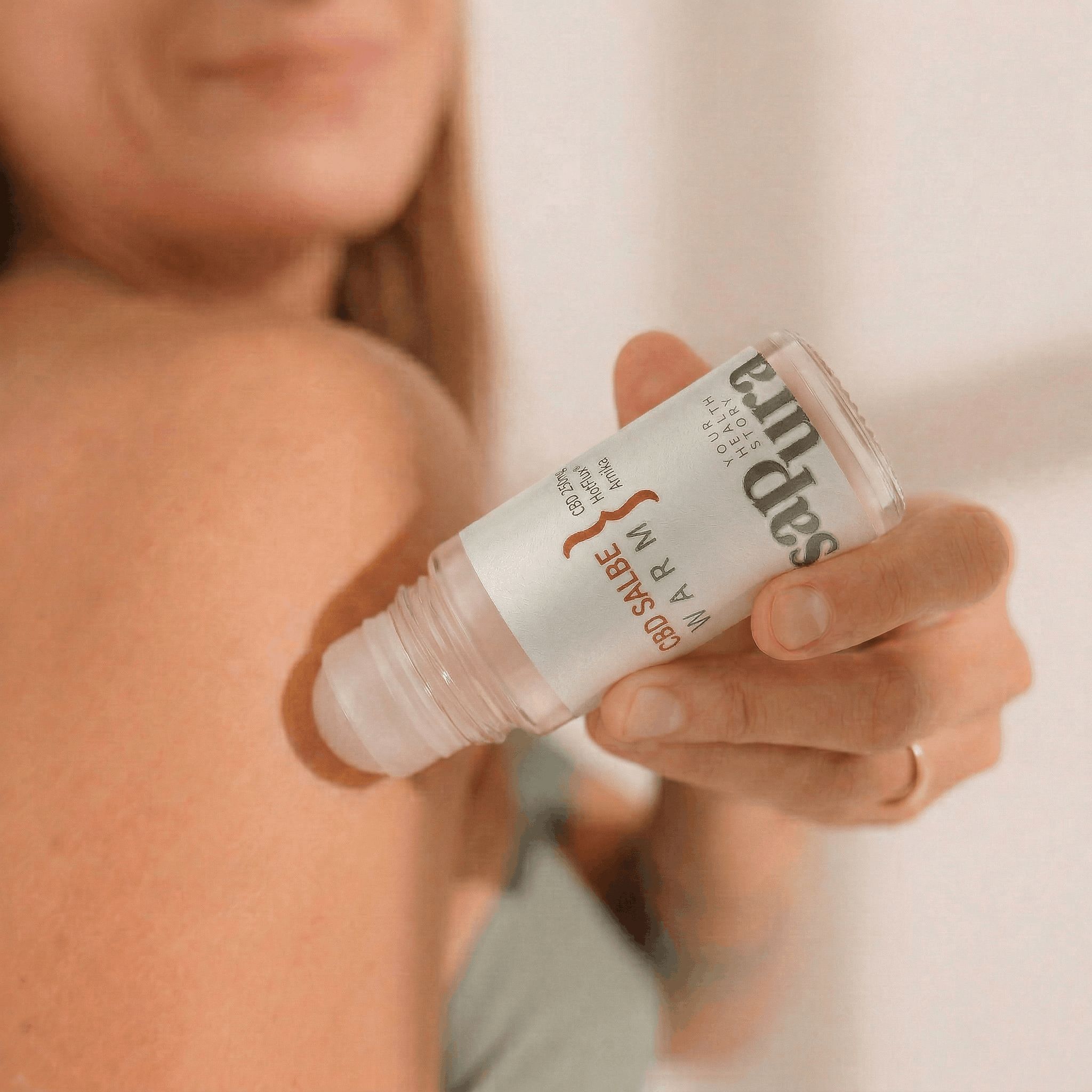 Hand hält eine CBD Salbe Warm Roll-On Flasche am Rücken einer Person. Der Applikator ist auf die Haut gerichtet.