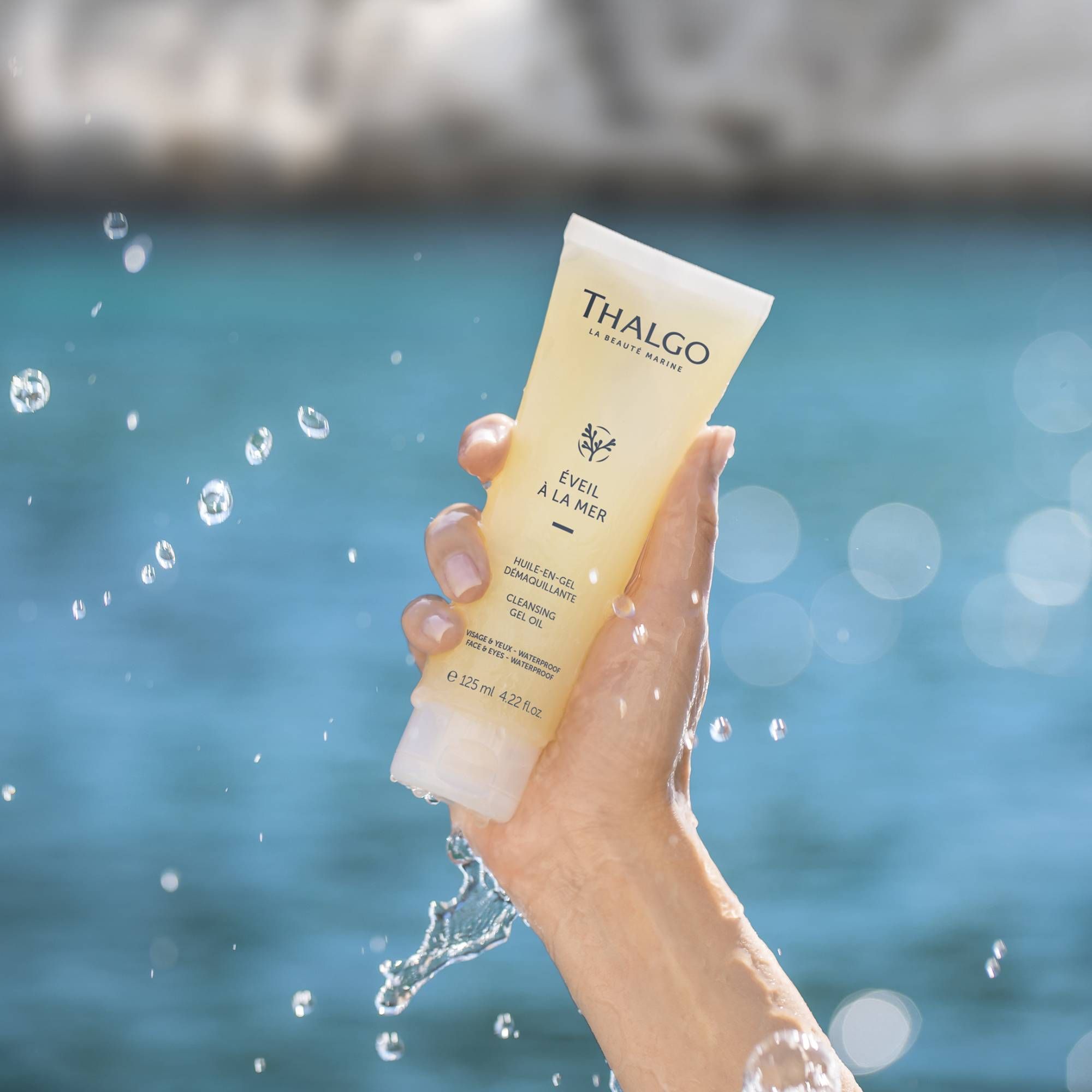 Hand hält Tube THALGO Reinigungsöl-Gel. Wasser spritzt. Logo und Produktname sichtbar.