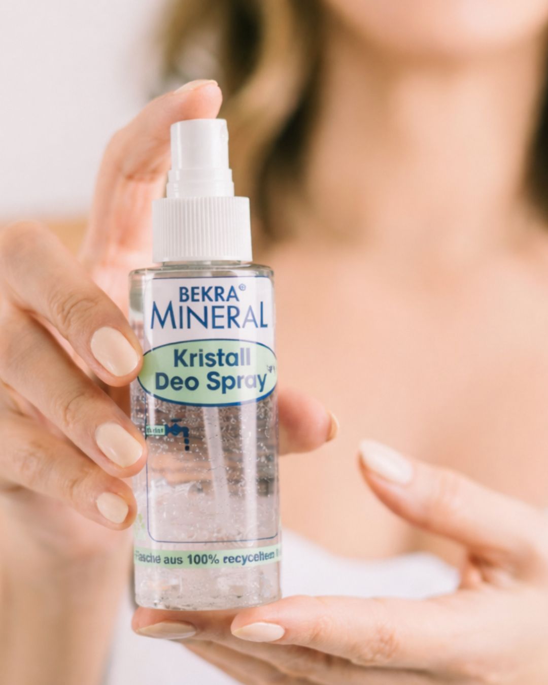 Hände halten eine Flasche BEKRA Mineral Kristall Deo Spray. Der Sprühkopf ist weiß.