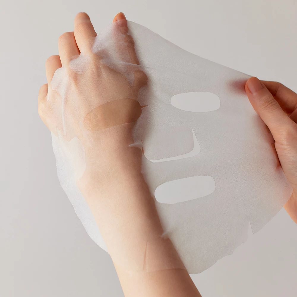 Hände halten eine transparente Gesichtsmaske. Die Maske hat Aussparungen für Augen, Nase und Mund.