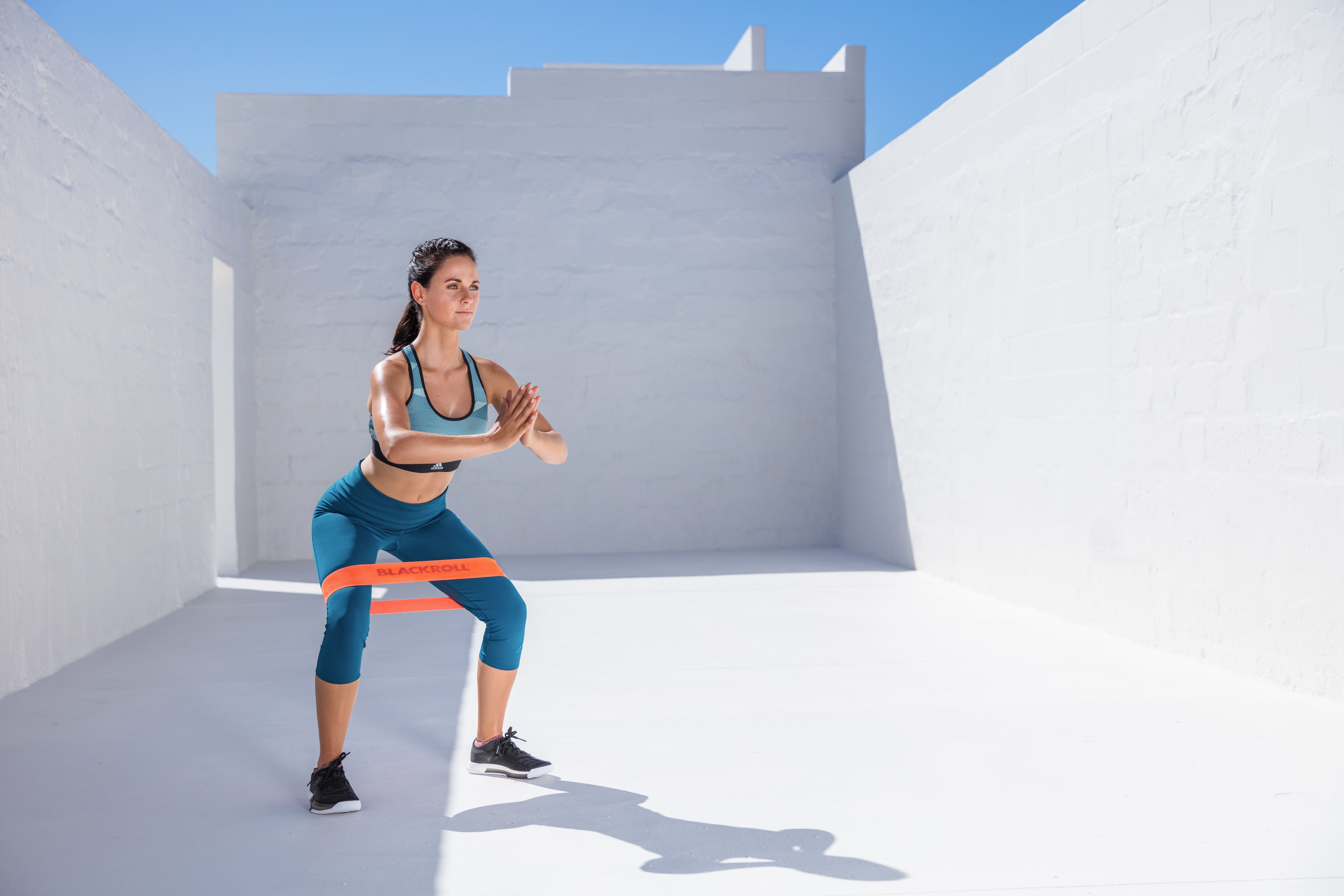 Frau macht Kniebeugen mit orangefarbenem Loop Band um die Oberschenkel. Blaue Sportkleidung.