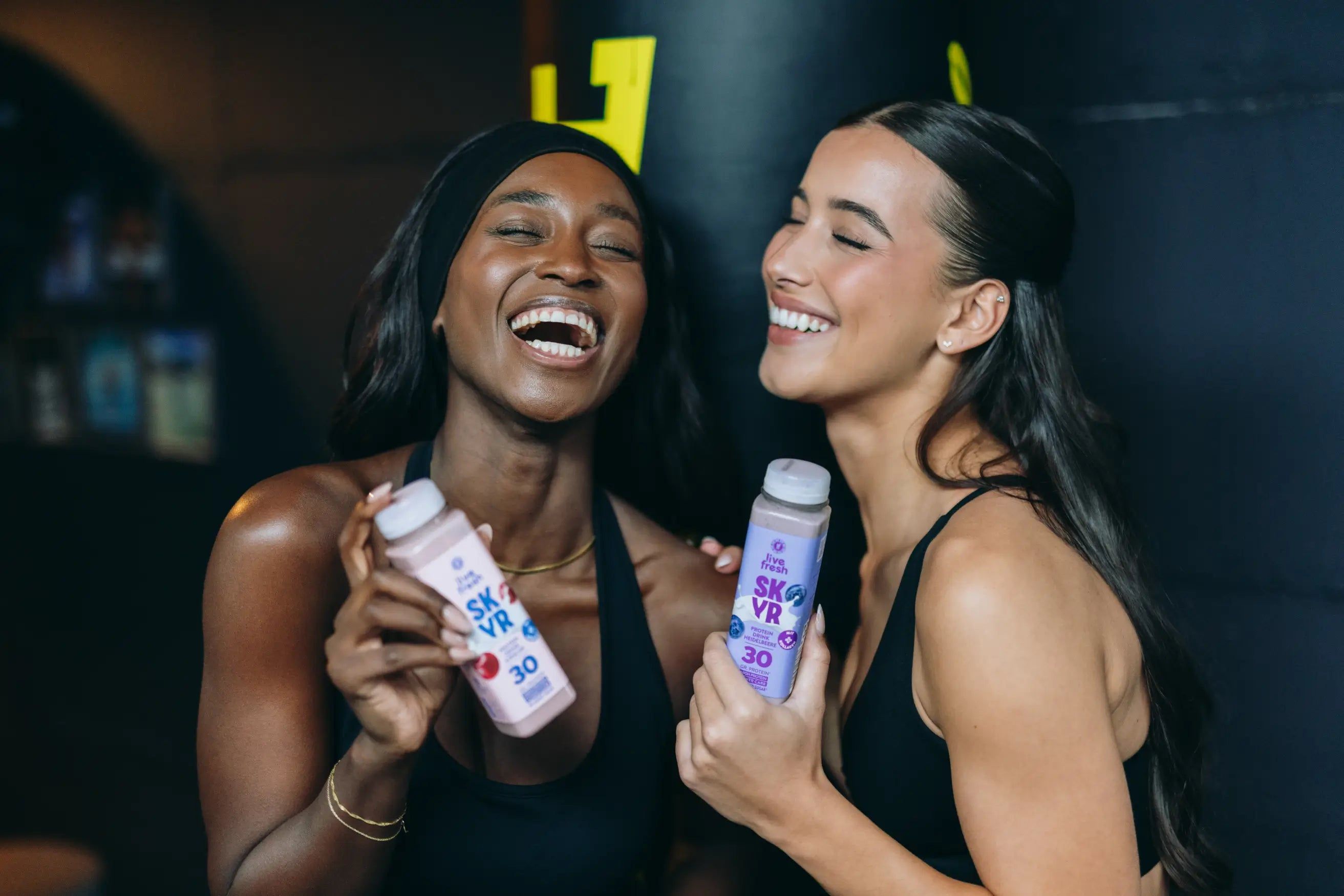 Zwei Frauen lachen und halten Protein-Drinks. Eine Frau hält einen Beeren-Skyr-Drink, die andere einen Blaubeer-Skyr-Drink.