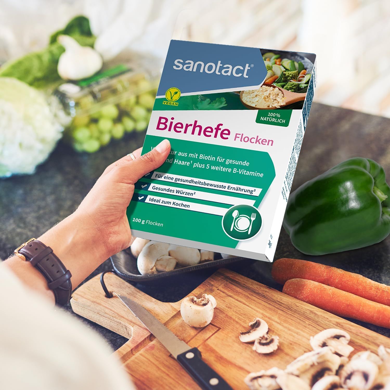 Person hält eine Packung Bierhefe Flocken. Sanotact-Logo. Text: 100 g Flocken. Auf einem Tisch mit Gemüse und Küchenutensilien.
