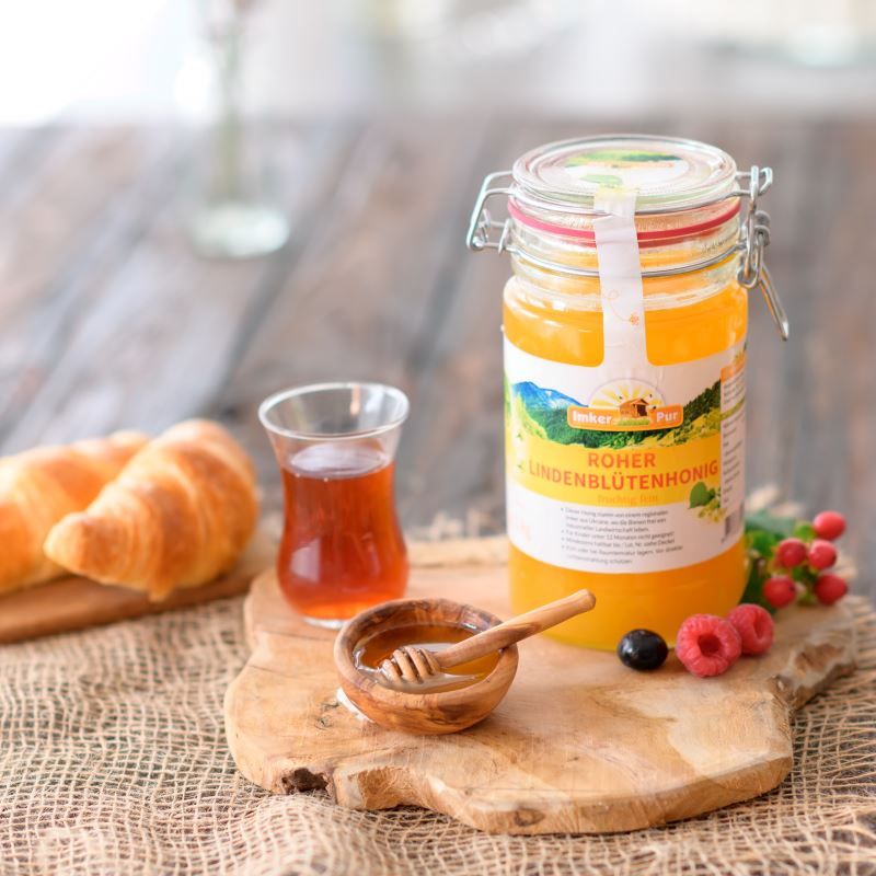 Honigglas mit Etikett, neben Glas mit Honig, Croissants, Holzbrett mit Beeren. Honig im Glas sichtbar.