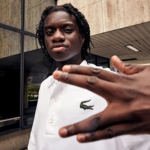 Ein junger Mann mit Zöpfen trägt ein weißes Polohemd mit grünem Krokodil-Logo. Im Hintergrund ein Gebäude.