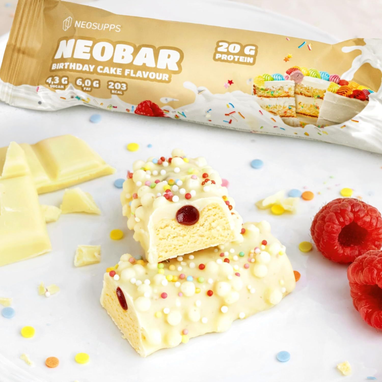 Zwei aufgeschnittene NeoBar-Stücke mit weißer Schokolade, Himbeeren und Streuseln. Daneben Himbeeren und Schokoladenstücke.
