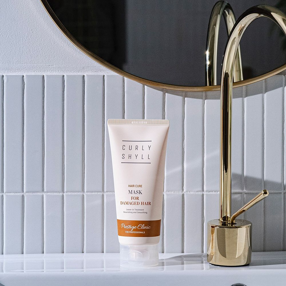 Beige Tube auf Waschbecken. Aufschrift: CURLY SHYLL, Hair Cure Mask for Damaged Hair. Prestige Clinic. Goldfarbene Armatur und Spiegel.