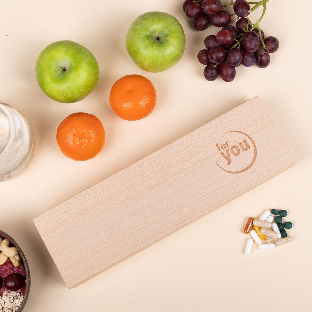 Geschlossene Tablettenbox aus Holz mit 'for you' Logo. Daneben Obst: Äpfel, Mandarinen, Trauben. Einige Tabletten daneben.