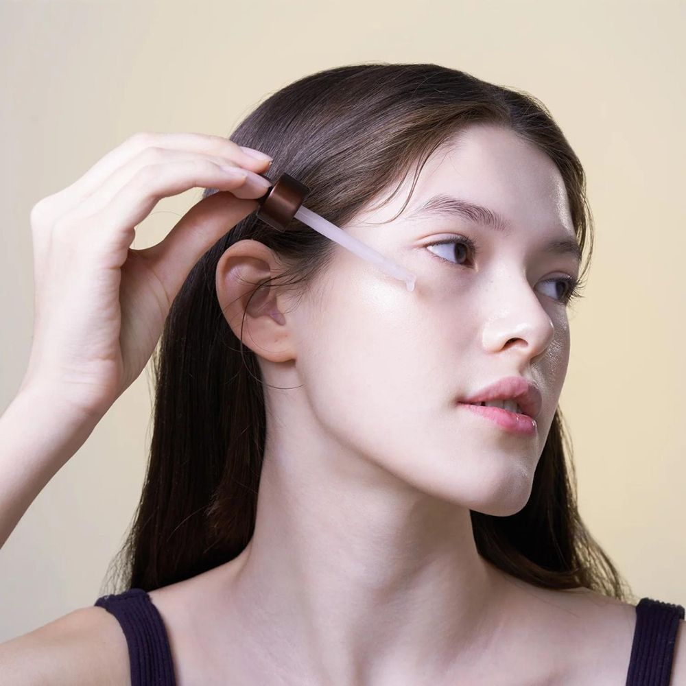Junge Frau mit langen Haaren, die mit einer Pipette Serum auf ihr Gesicht aufträgt. Helle Haut, Blick zur Seite.