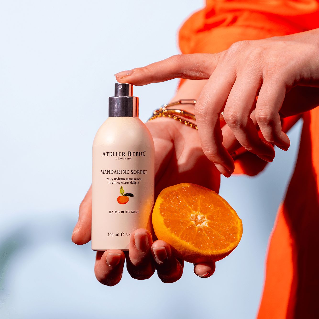 Hände halten eine Sprühflasche und eine Orangenhälfte. Auf der Flasche steht "MANDARINE SORBET" und "HAIR & BODY MIST".