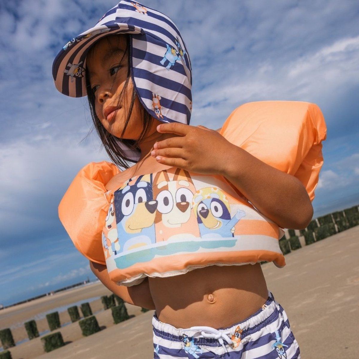 Kind am Strand mit Schwimmhilfe und Hut. Orangefarbene Arme, Brustteil mit Bluey-Motiv, blau-weiß gestreifter Hut.