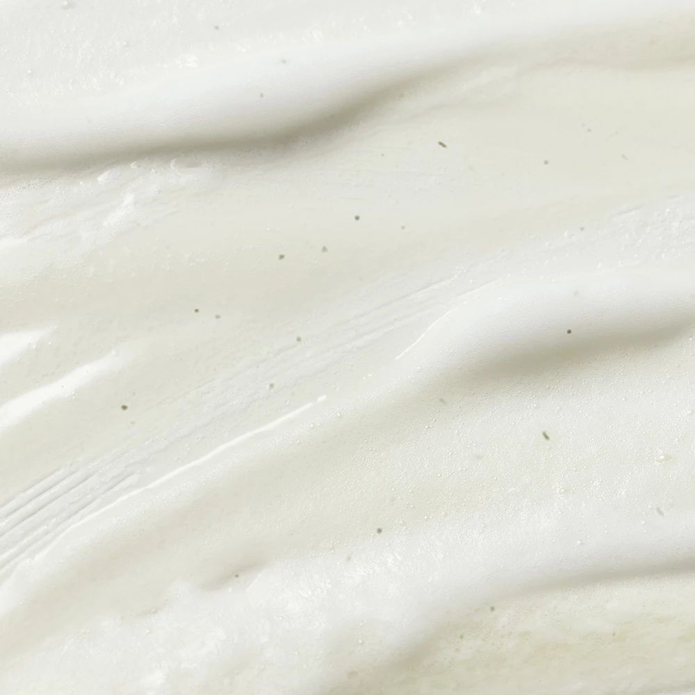 Nahaufnahme von weißer, cremiger Substanz. Kleine grüne Partikel sind sichtbar. Textur ist glatt.