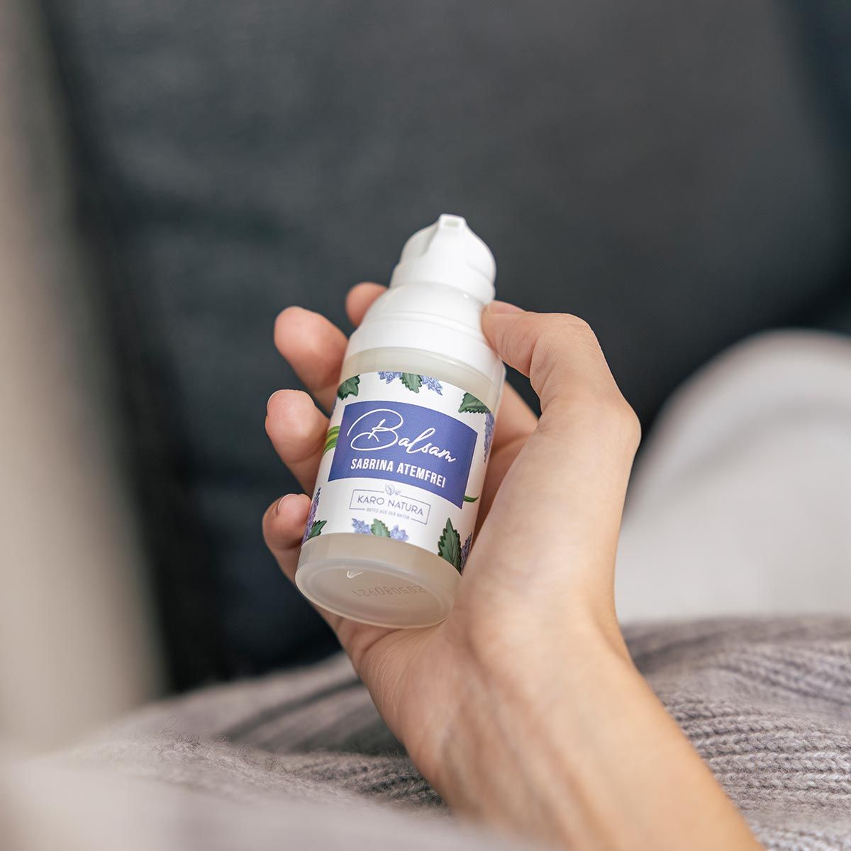 Person hält Balsam-Spender. Aufschrift: Balsam Sabrina Atemfrei. Karo Natura Logo. Graue Decke im Hintergrund.