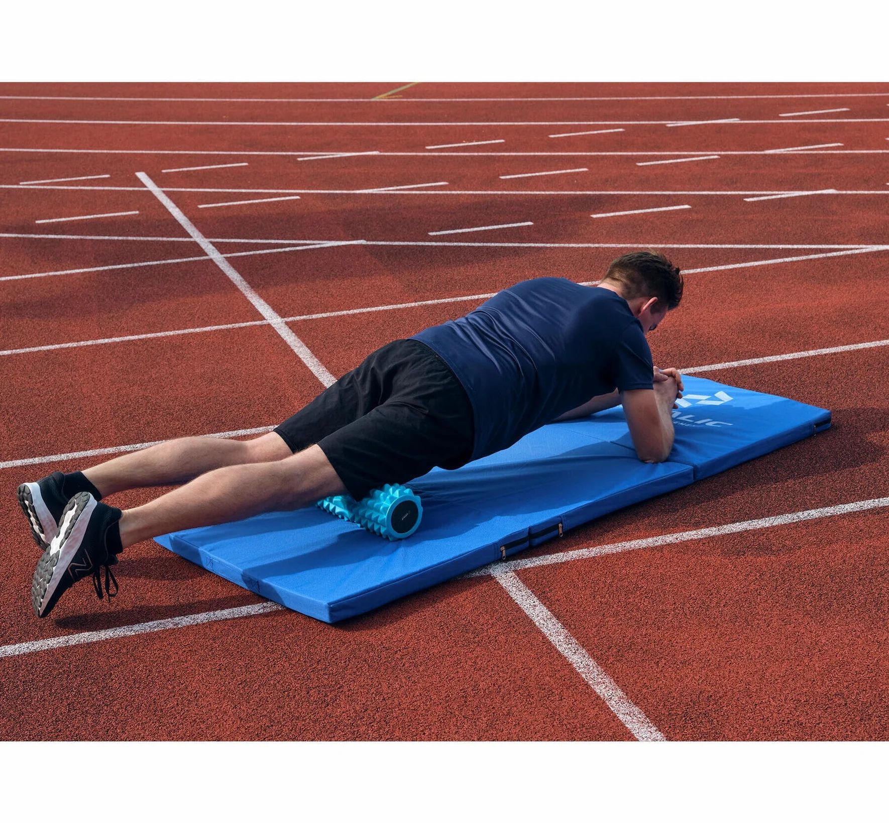 Mann trainiert auf blauer Matte mit Faszienrolle. Auf roter Laufbahn.