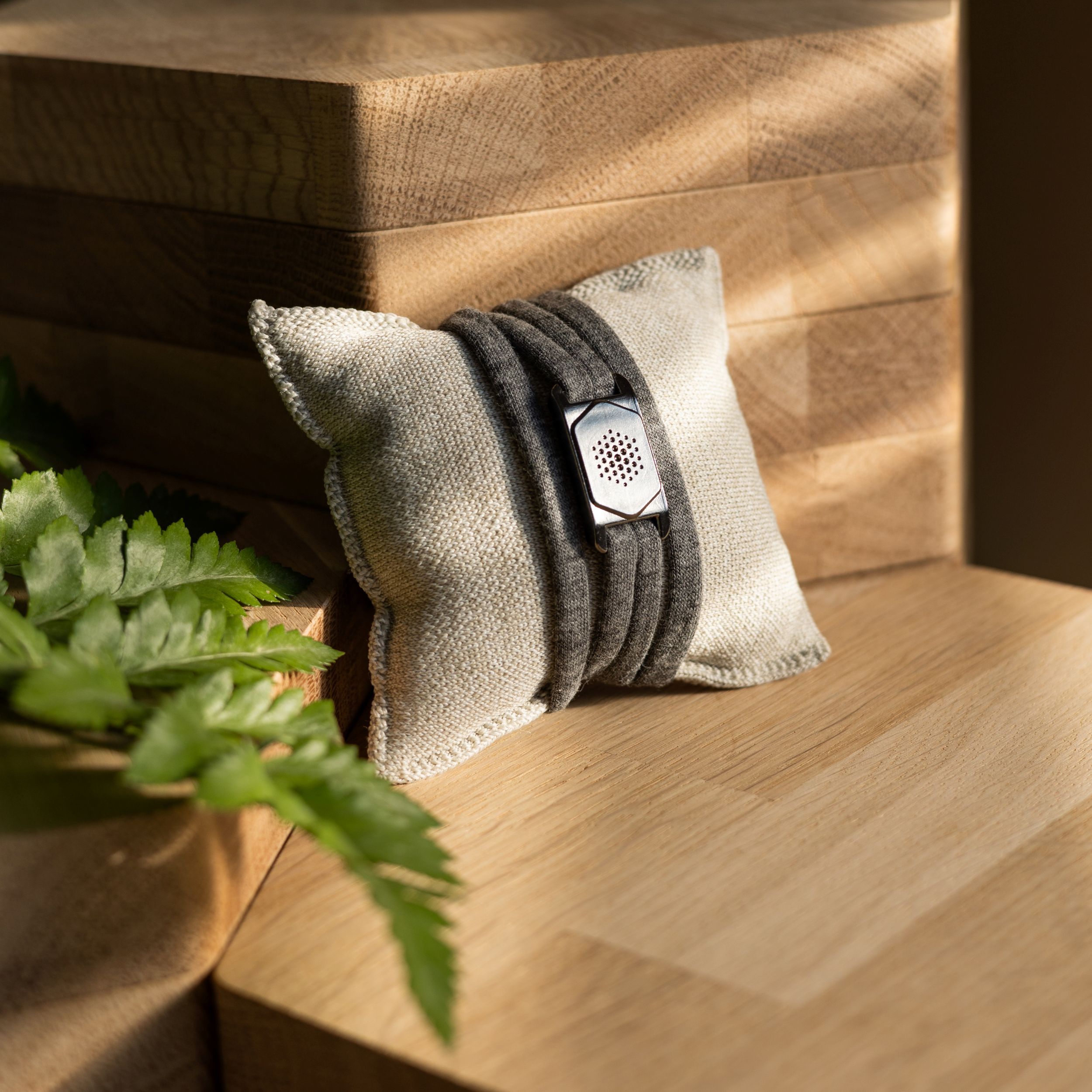 Graues Armband auf einem kleinen, quadratischen Kissen. Das Armband hat ein dunkles, rechteckiges Element. Holzuntergrund und grüne Blätter.