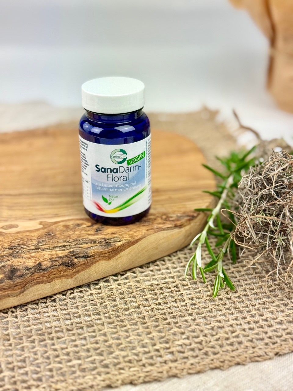 Blaue Flasche SanaDarm-Floral auf Holzbrett. Vegan-Siegel. Etikett mit Produktinformationen. Rosmarinzweig.