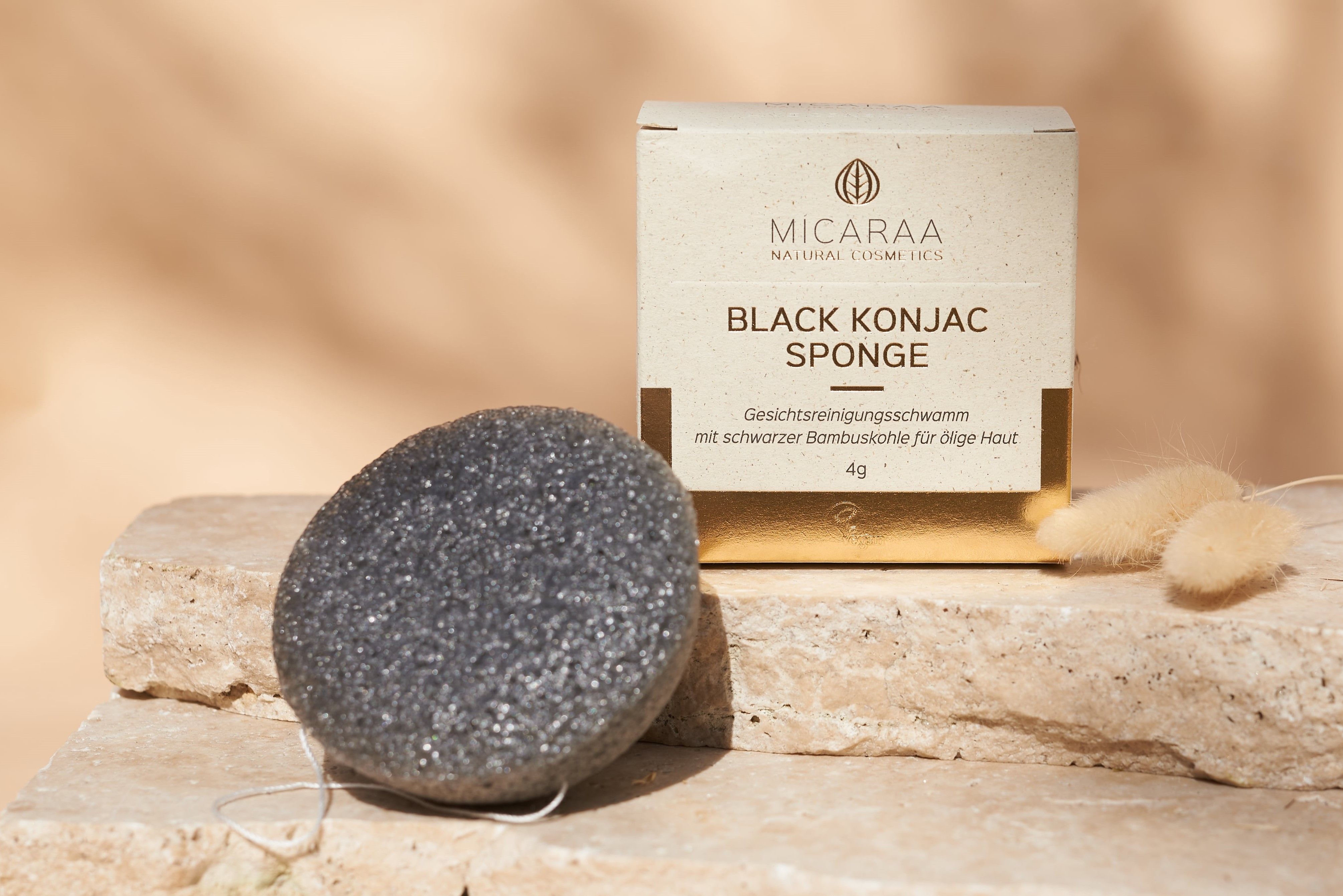 Grauer, runder Schwamm und Verpackung auf Stein. Verpackung mit Logo und Text: BLACK KONJAC SPONGE.