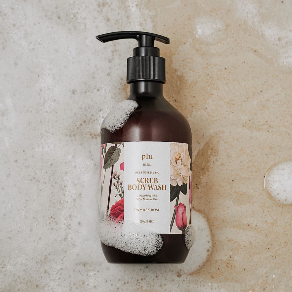 Braune Flasche mit schwarzem Pumpverschluss, umgeben von Schaum. Etikett mit floralem Design und Schriftzug: Scrub Body Wash, Damask Rose. Marke: plu.