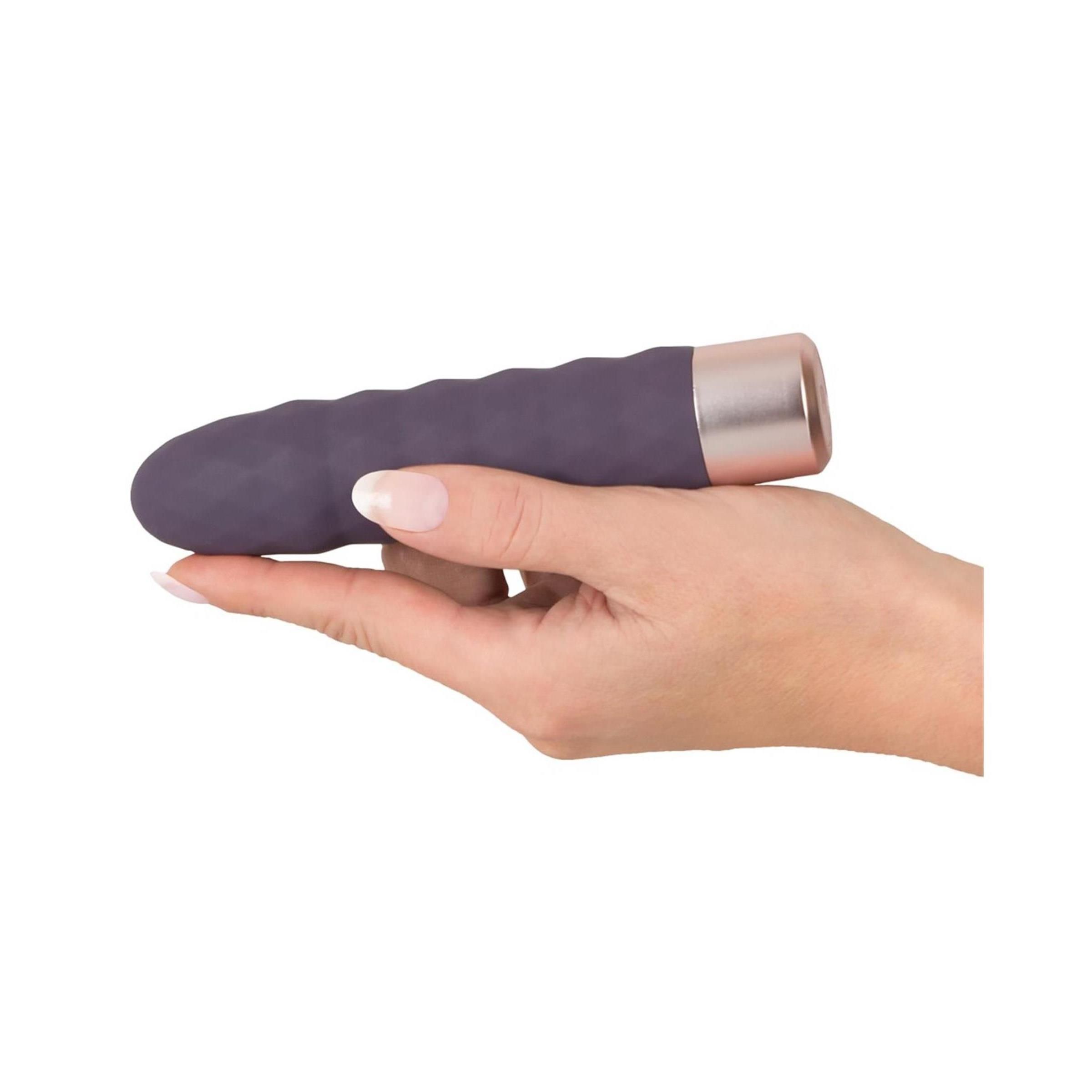 Dunkelvioletter Vibrator in Hand gehalten. Goldfarbener Sockel. Geriffelte Oberfläche. Zylindrische Form.