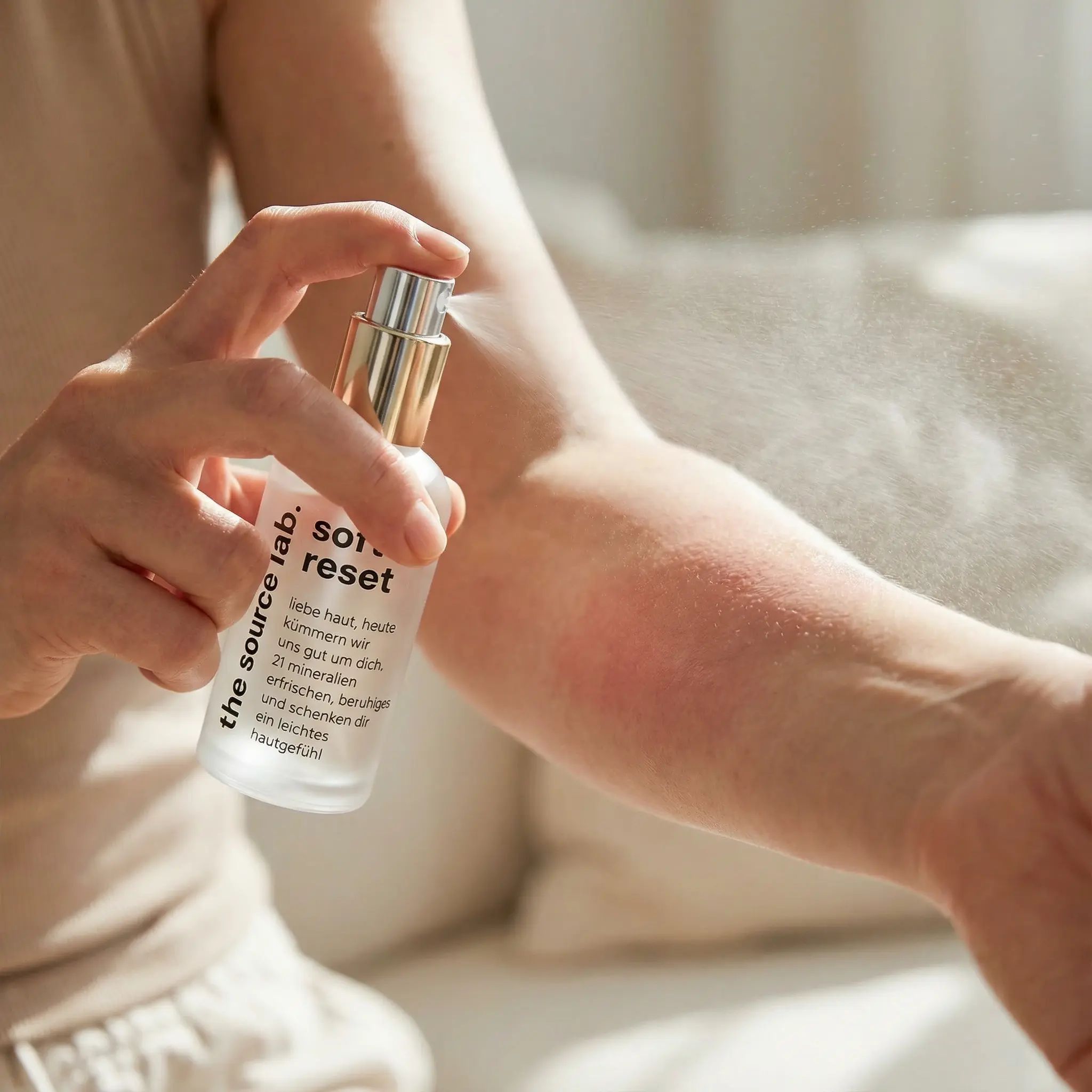 Person sprüht Hautspray auf Arm. Rötung sichtbar. Produkt: "soft reset" von the source lab. Goldener Sprühkopf, transparente Flasche.