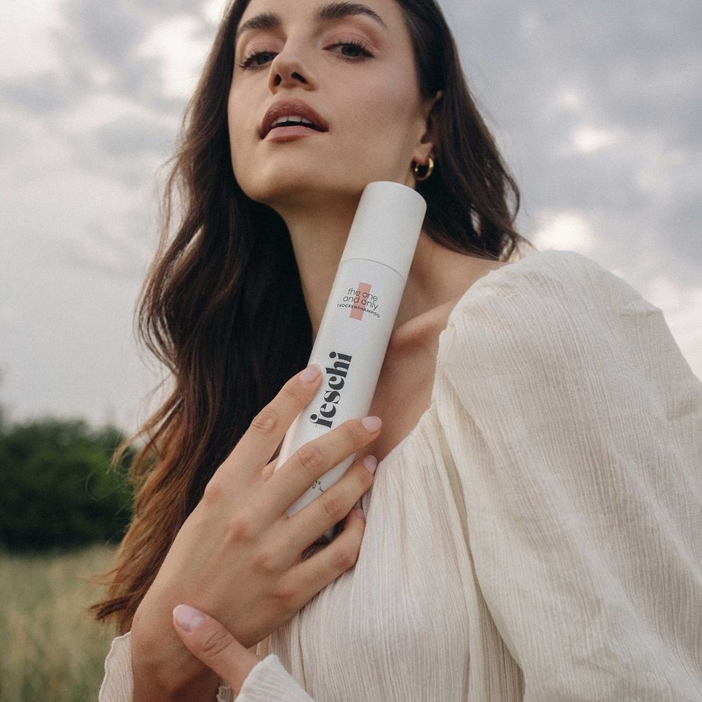 Frau mit weißem Kleid hält das weiße Trockenshampoo-Spray. Das Produkt wird in der Hand gehalten.