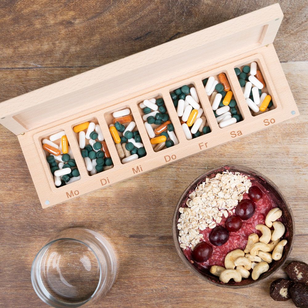 Geöffnete Tablettenbox aus Holz mit Tabletten in sieben Fächern. Beschriftung mit Wochentagen. Daneben eine Schüssel mit Essen.