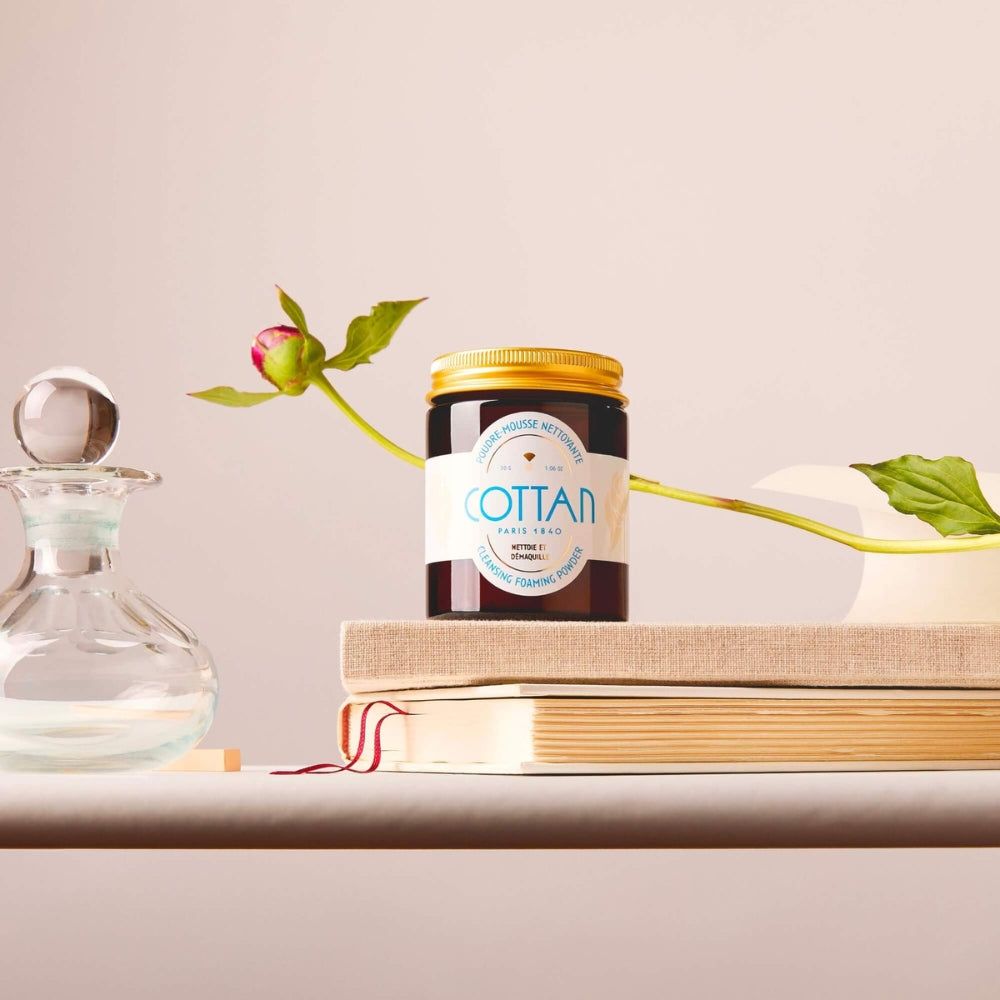 Braune Glasdose mit goldenem Deckel. Aufschrift "COTTAN". Auf Büchern, neben einer Glasflasche und einer Blüte.