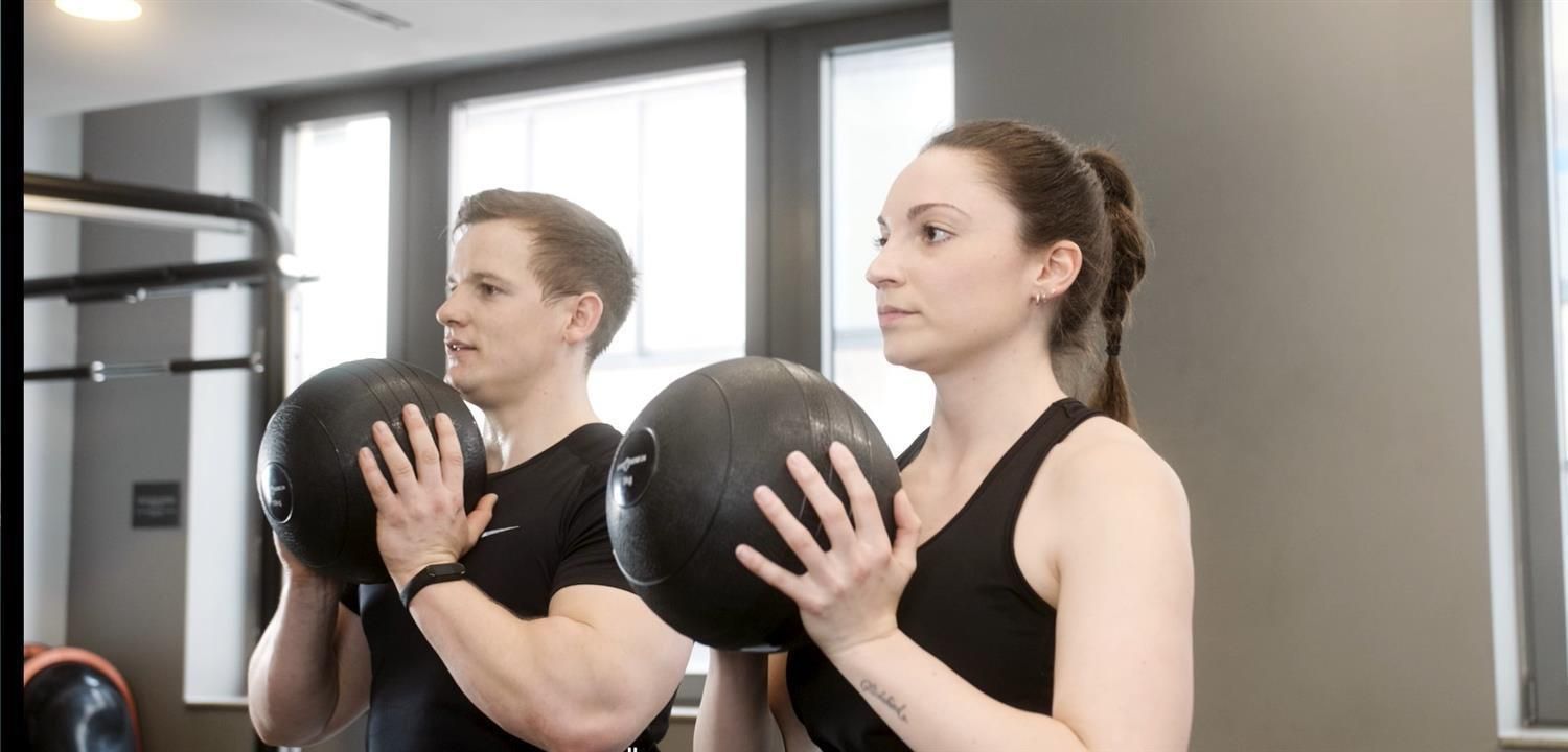 Zwei Personen halten jeweils einen schwarzen Slamball. Fitnessstudio-Umgebung.