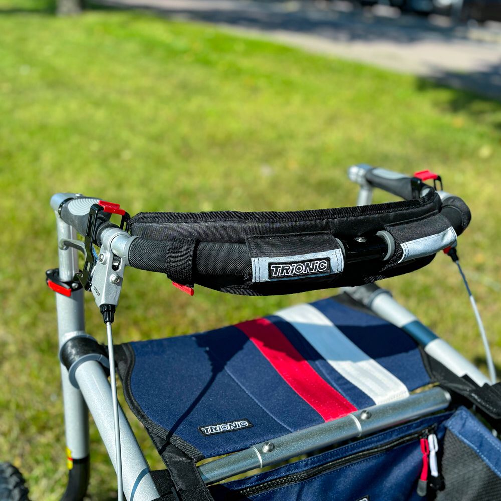 Schwarzes Rückenpolster mit Trionic-Logo am Rollator. Rote Akzente. Blaue Sitzfläche mit roten und weißen Streifen. Grüne Wiese im Hintergrund.