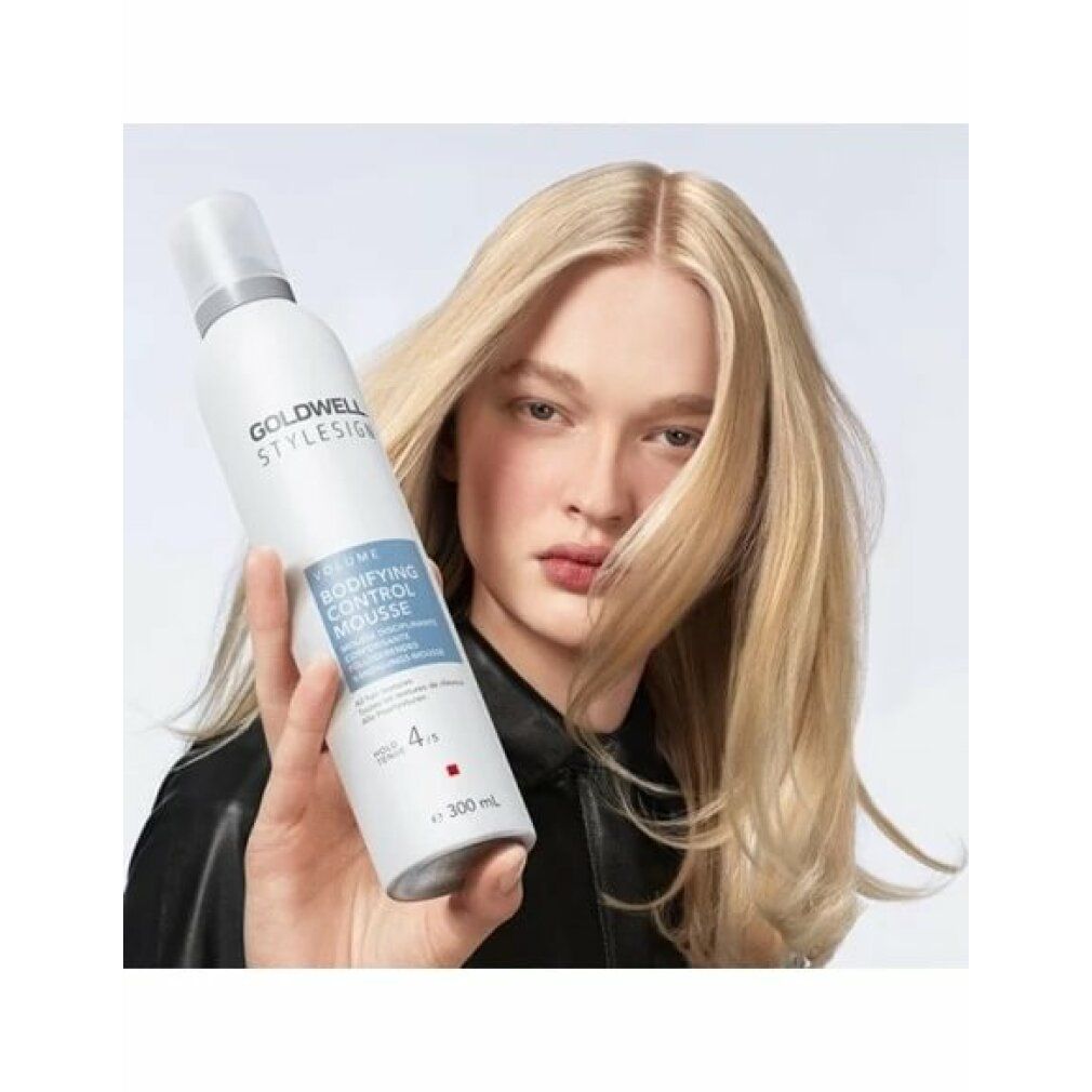 Frau hält Goldwell Stylesign Volume Bodifying Control Mousse. Blondes Haar. Schwarze Jacke. Produktetikett sichtbar.