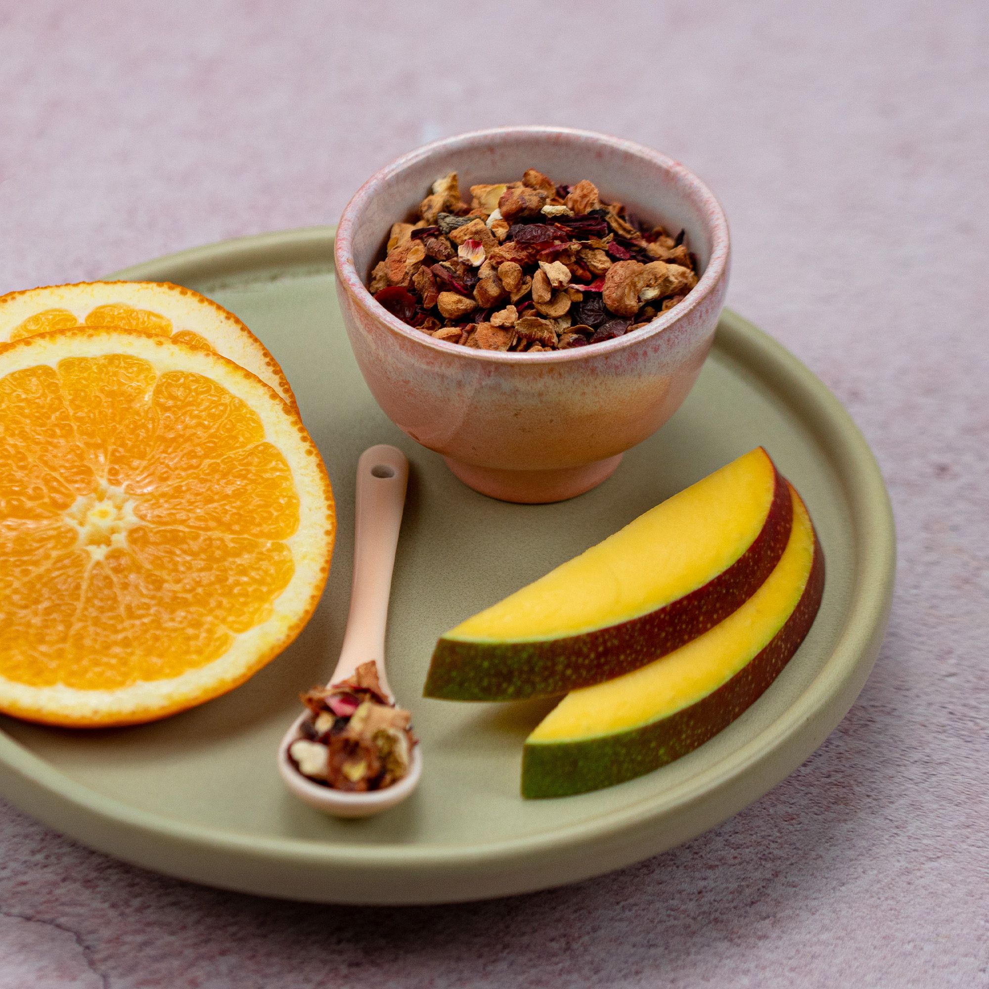 Schale mit Teemischung, daneben Löffel mit Tee, Mango- und Orangenscheiben auf Untertasse.