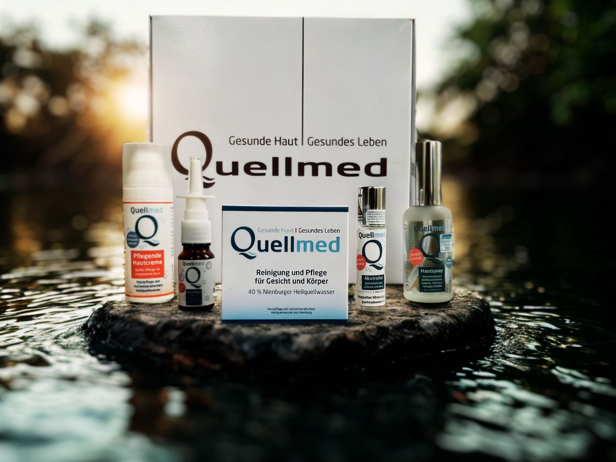 Quellmed-Produkte auf einem Stein vor Wasser. Enthalten Cremes, Sprays und eine Box. Aufschrift: Quellmed, Gesunde Haut, Gesundes Leben.