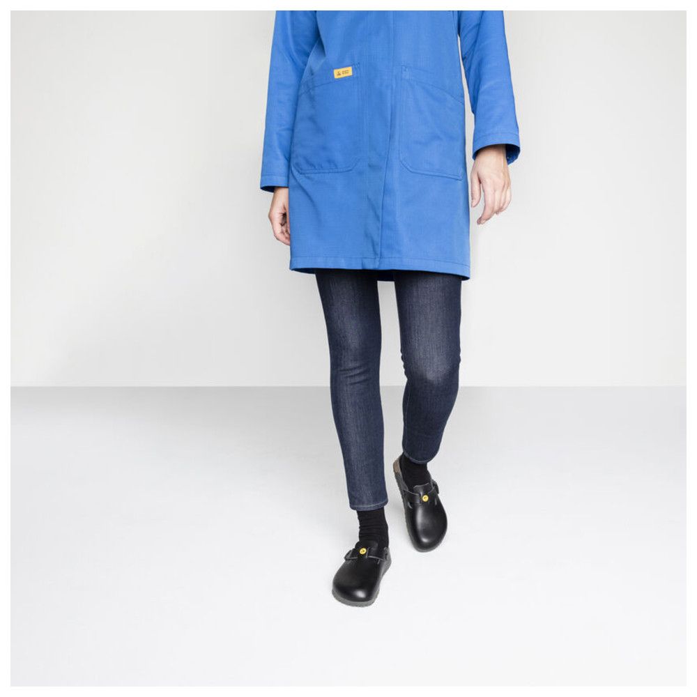 Person trägt schwarze Pantoletten. Blaue Arbeitsjacke und Jeans. Gelbes Logo auf dem Schuh.