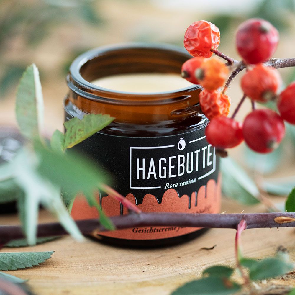 Braunes Glas mit schwarzem Etikett HAGEBUTTE auf Holz. Rote Hagebutten und Blätter im Hintergrund. ZEROX Logo.