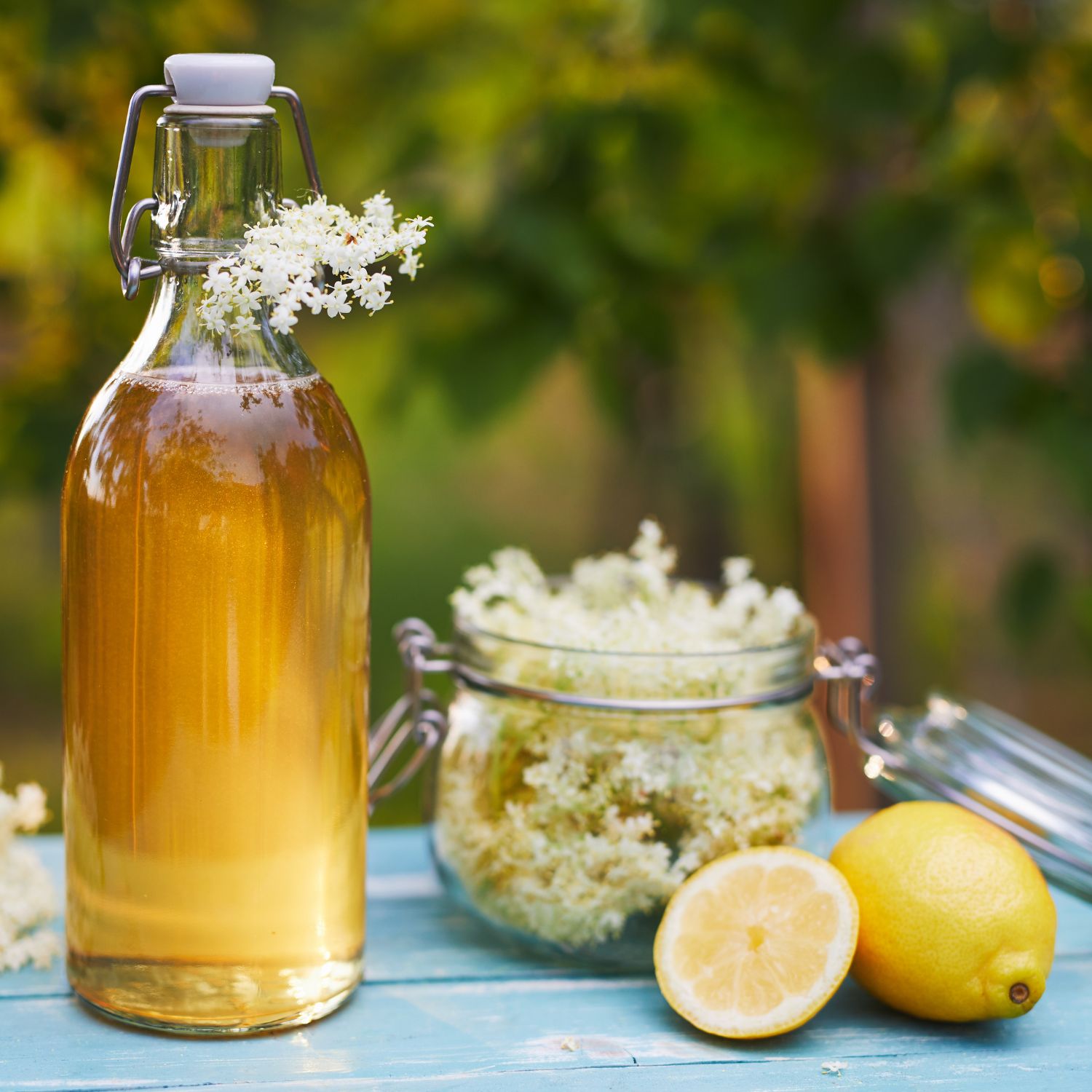 Glasflasche mit Flüssigkeit, daneben Glasbehälter mit Blüten. Zitronen und Blüten auf blauem Tisch. Hintergrund: Garten.