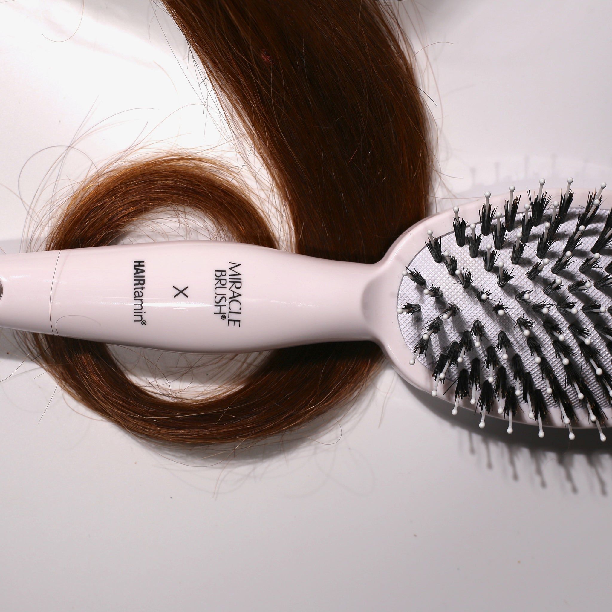 Weiße Bürste mit schwarzen Borsten und einem Haarstrang. Aufschrift MIRACLE BRUSH X HAIRtamin.