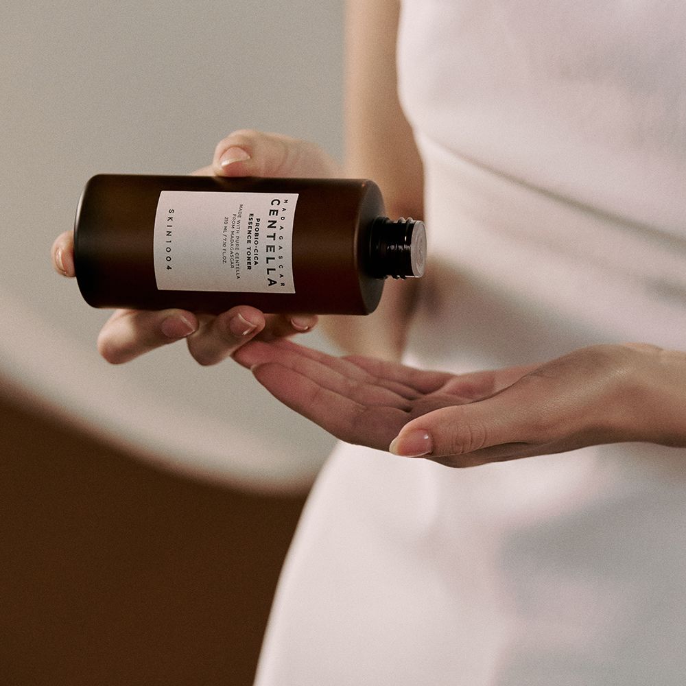 Person hält braune Flasche mit weißem Deckel. Aufkleber mit Text: CENTELLA, ESSENCE TONER, SKIN1004. Hände.