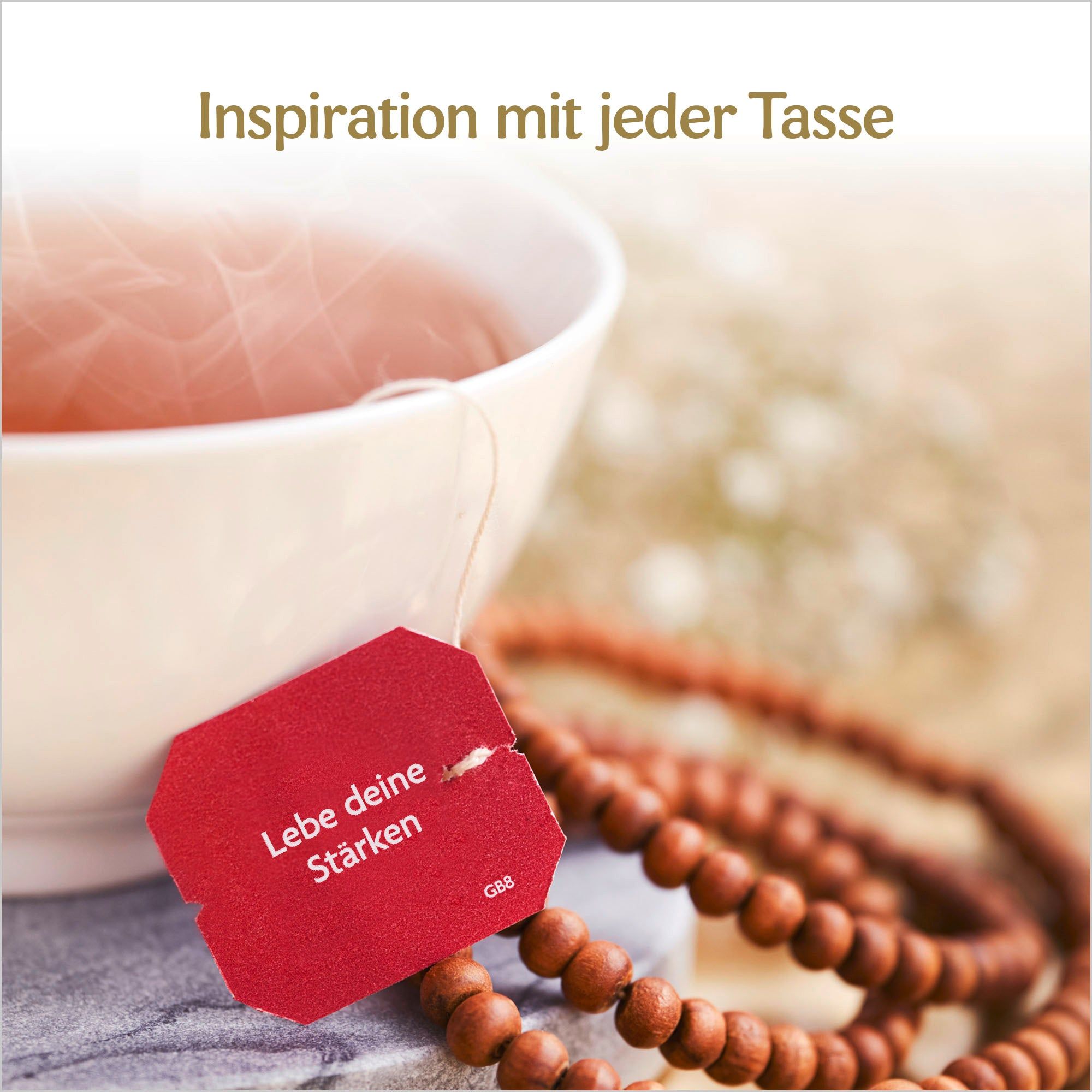 Tasse Tee mit rotem Teebeutel. Text: Lebe deine Stärken. Braune Gebetskette. Hintergrund unscharf.