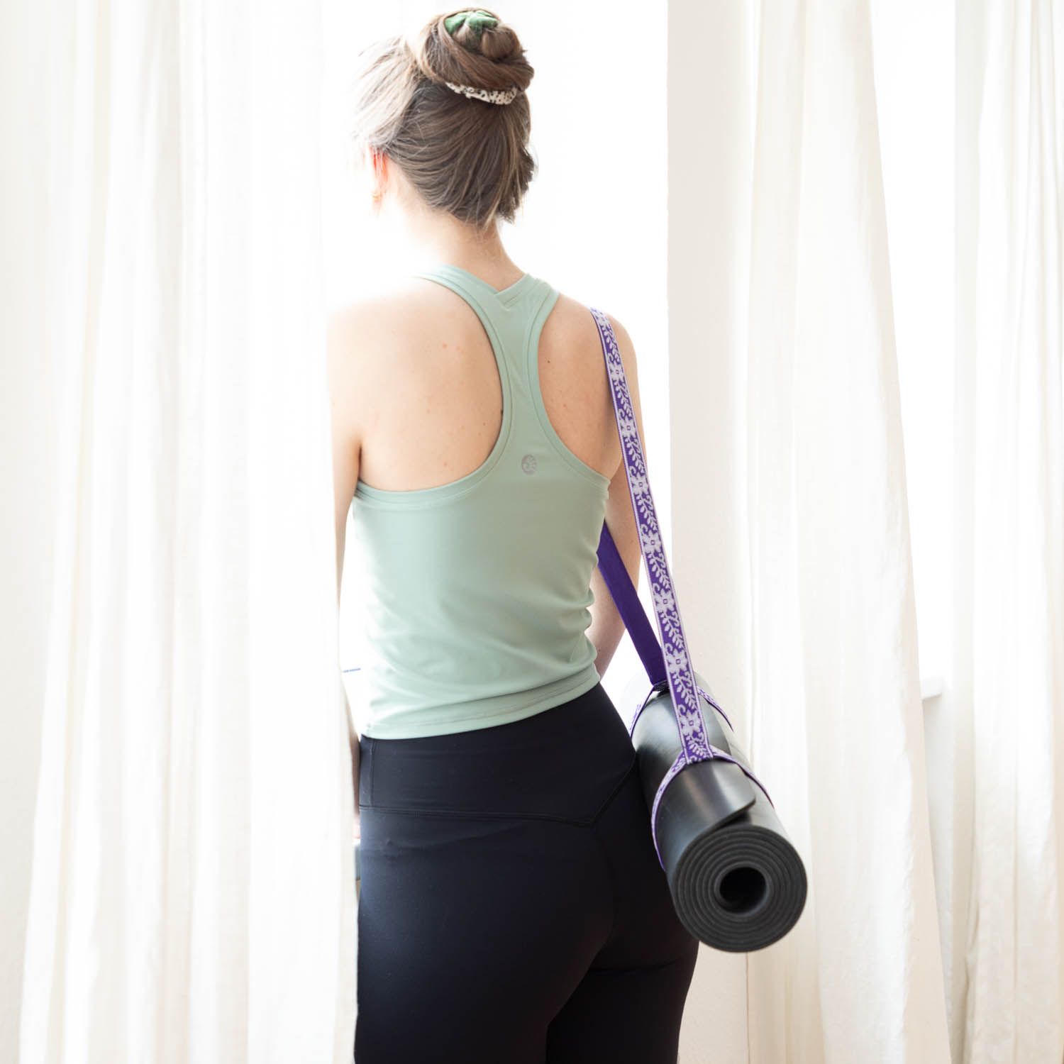 Person trägt Yogamatte mit lila Tragegurt. Matte gerollt. Frau steht am Fenster.