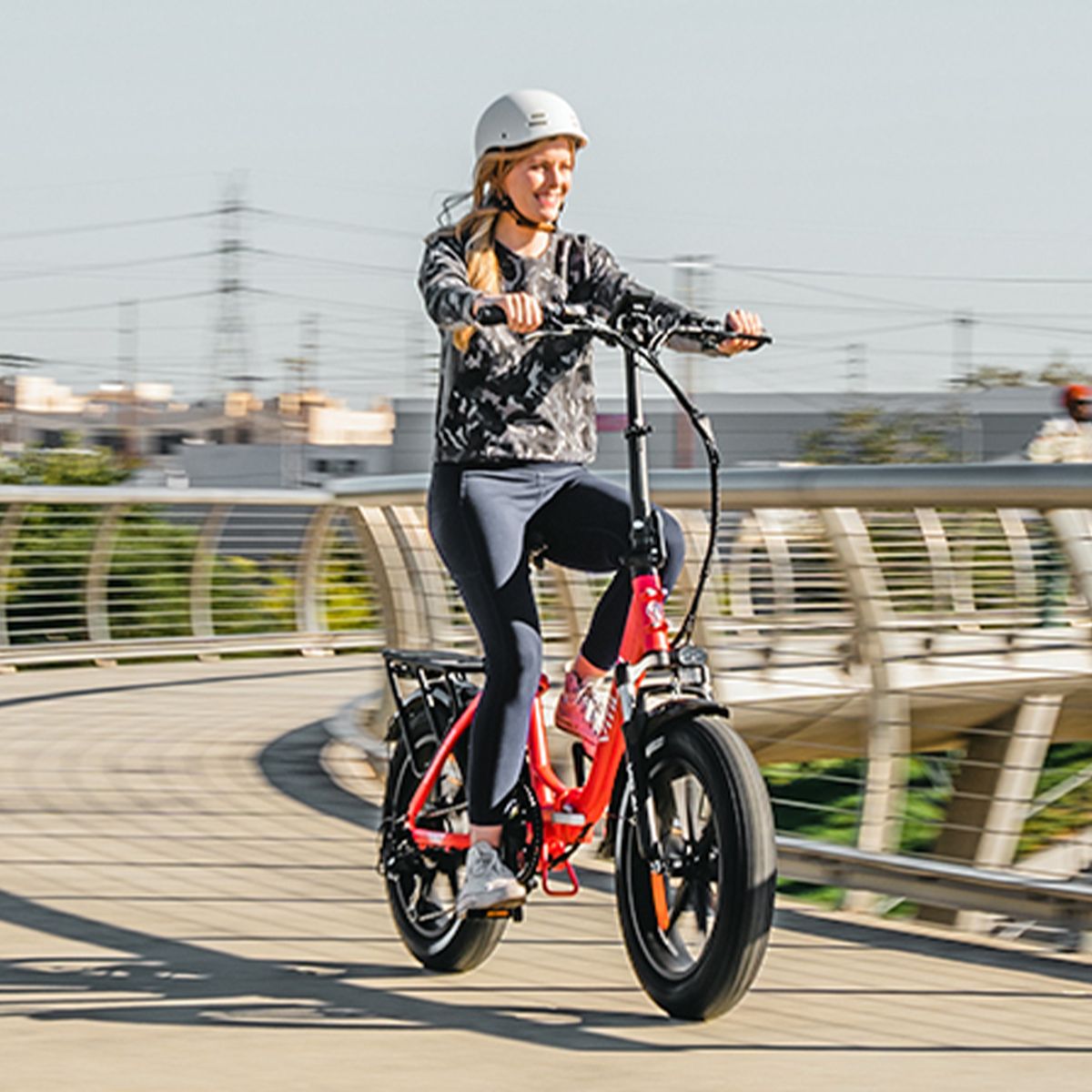 Person fährt auf rotem Elektrofahrrad. Frau mit Helm auf Brücke. Hintergrund: Gebäude und Himmel. Fahrrad mit schwarzen Reifen.