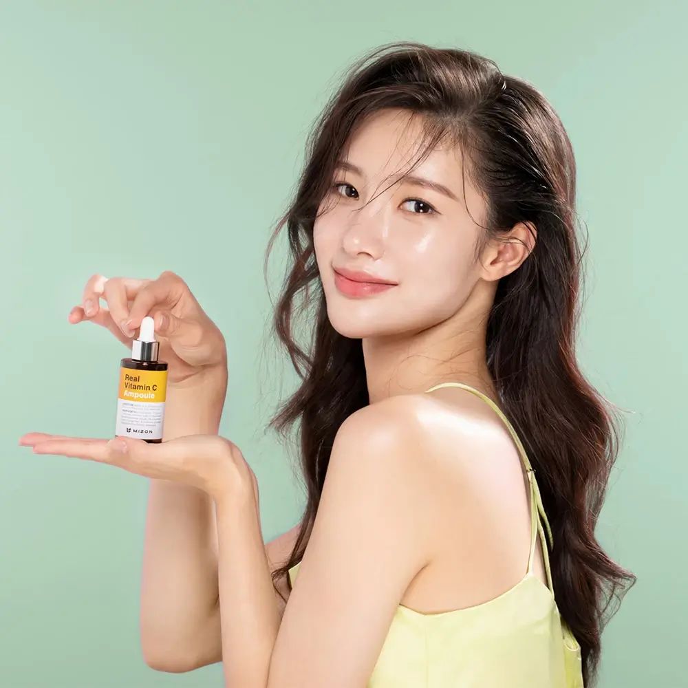 Frau hält eine kleine Flasche mit gelbem Etikett. Aufschrift: Real Vitamin C Ampoule. Grüner Hintergrund.