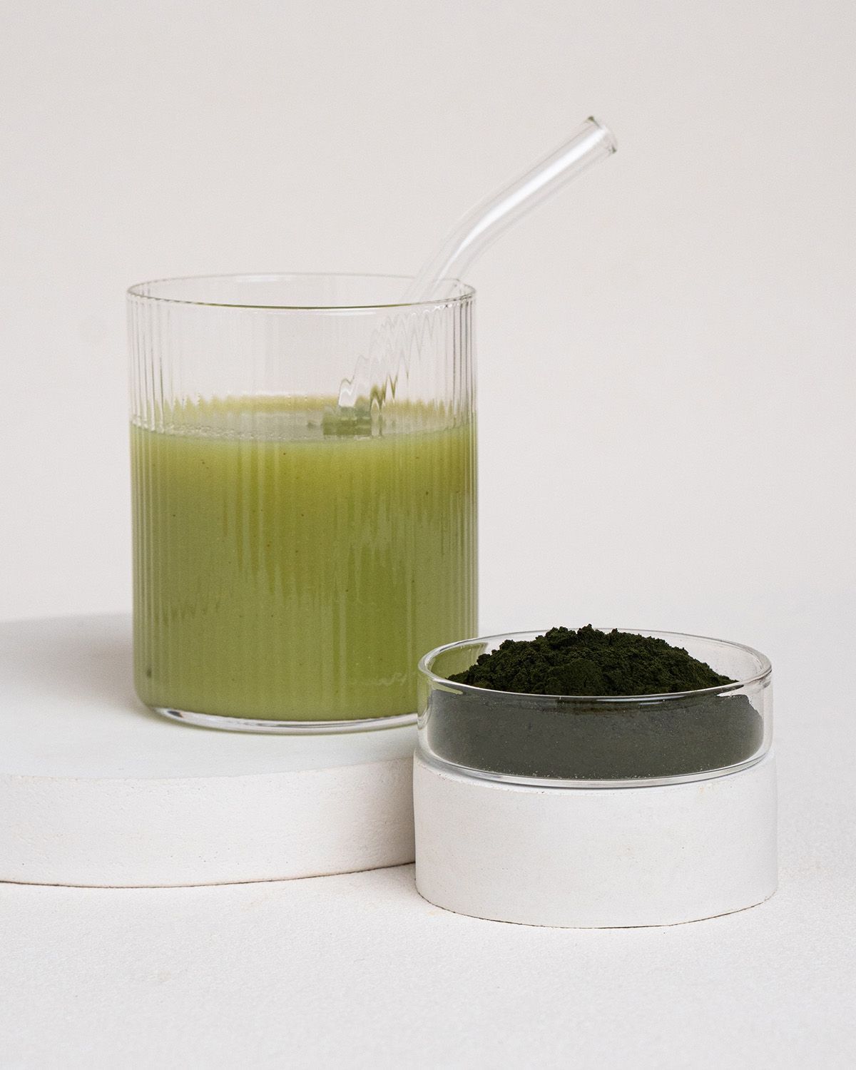 Glas mit grünem Getränk, daneben ein Behälter mit Chlorella-Pulver. Mit Trinkhalm. Auf weißem Podest.