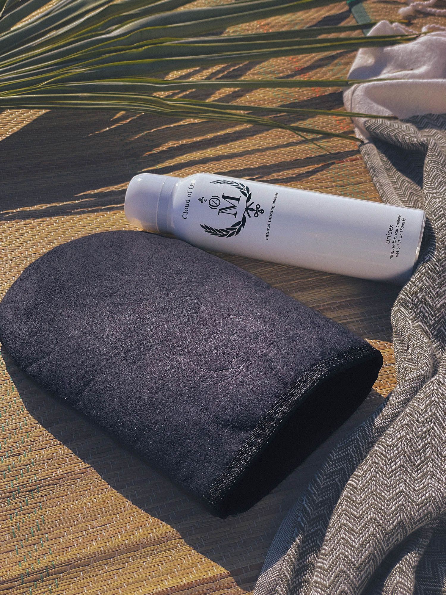 Weiße Sprühflasche mit schwarzem Logo und Schriftzug neben einem schwarzen Applikatorhandschuh. Aufschrift: Cloud of Oz, Natural Tanning Mousse.