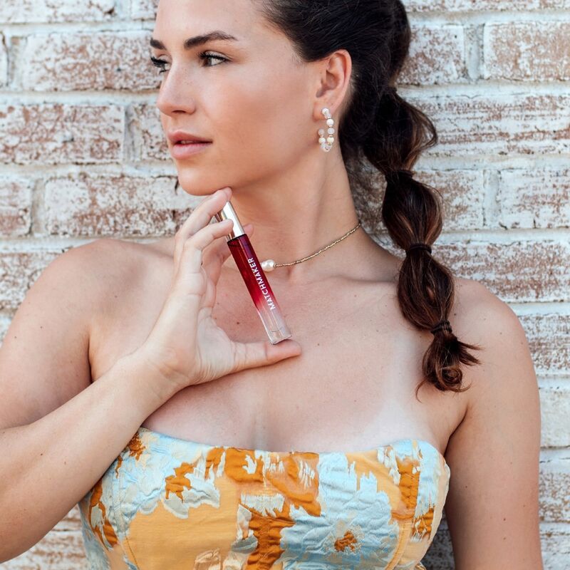 Klarer Flakon mit roter Flüssigkeit. Silberner Deckel. Aufschrift MATCHMAKER. Frau hält Flakon. Hintergrund: Ziegelmauer.