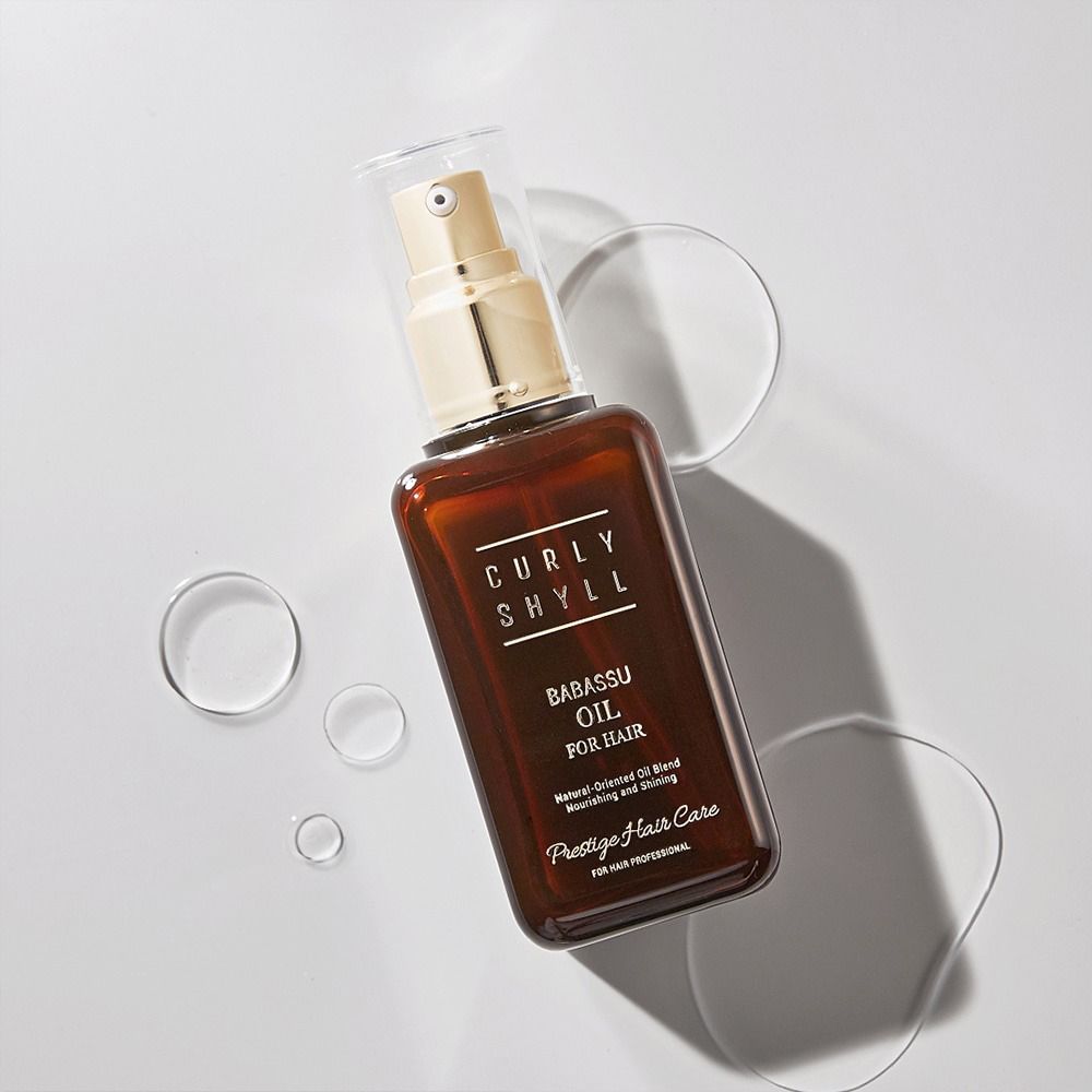 Braune Glasflasche mit goldfarbenem Sprühkopf. Aufschrift: CURLY SHYLL Babassu Oil for Hair. Umgeben von Wassertropfen.