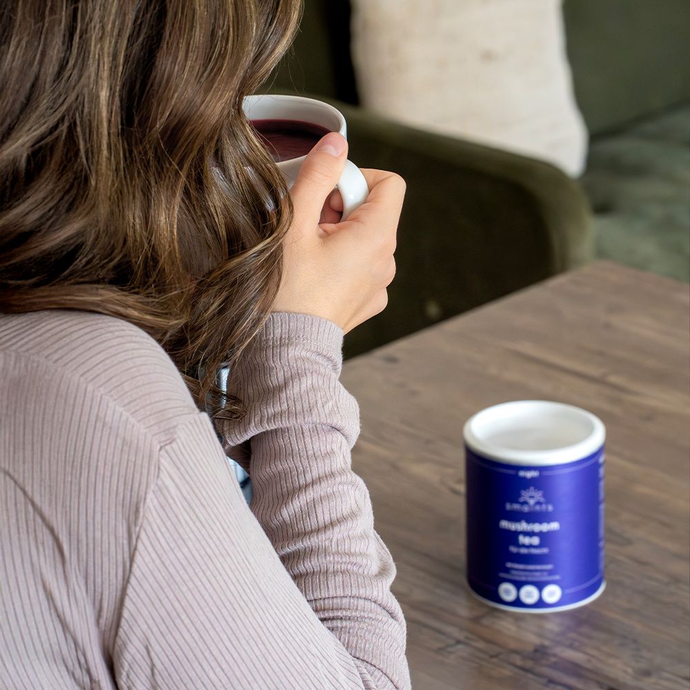 Person hält eine Tasse. Auf einem Tisch steht eine blaue Dose mit Text: Mushroom tea für die Nacht.