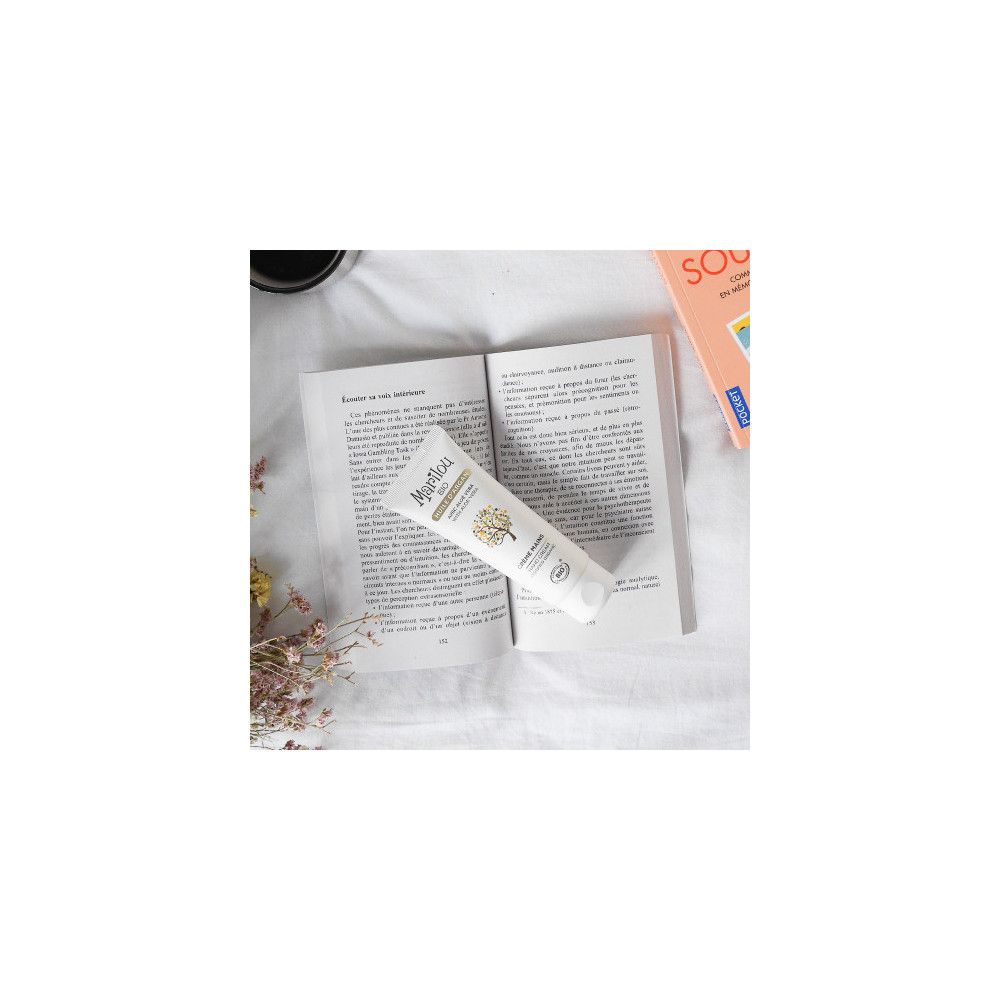 Weiße Tube Marilou Bio Handcreme auf offenem Buch. Logo und Produktbezeichnung sichtbar. Umgebung: Buch, Blumen.