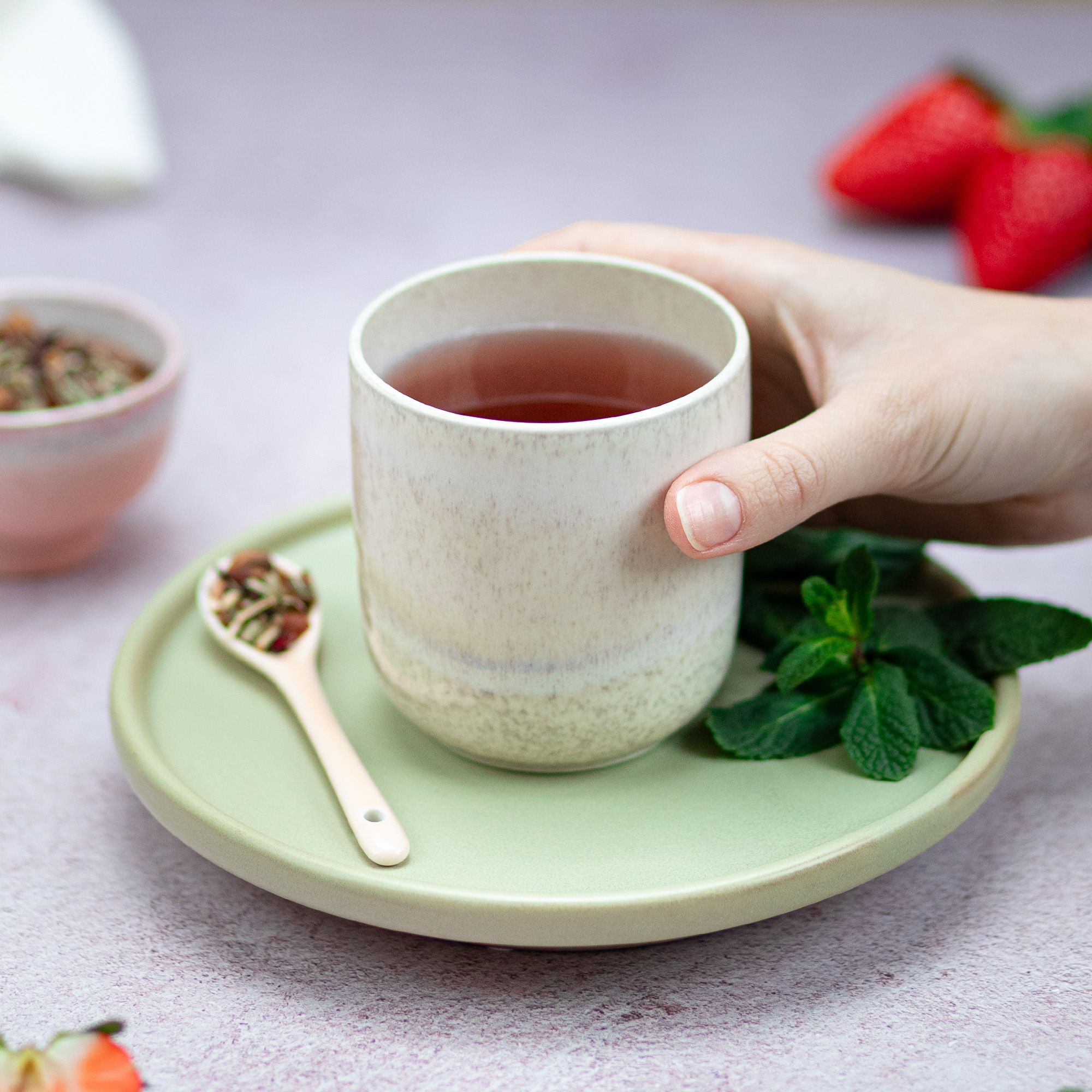 Tasse Tee, gehalten von Hand. Auf Untertasse mit Löffel und Minze. Erdbeeren im Hintergrund.