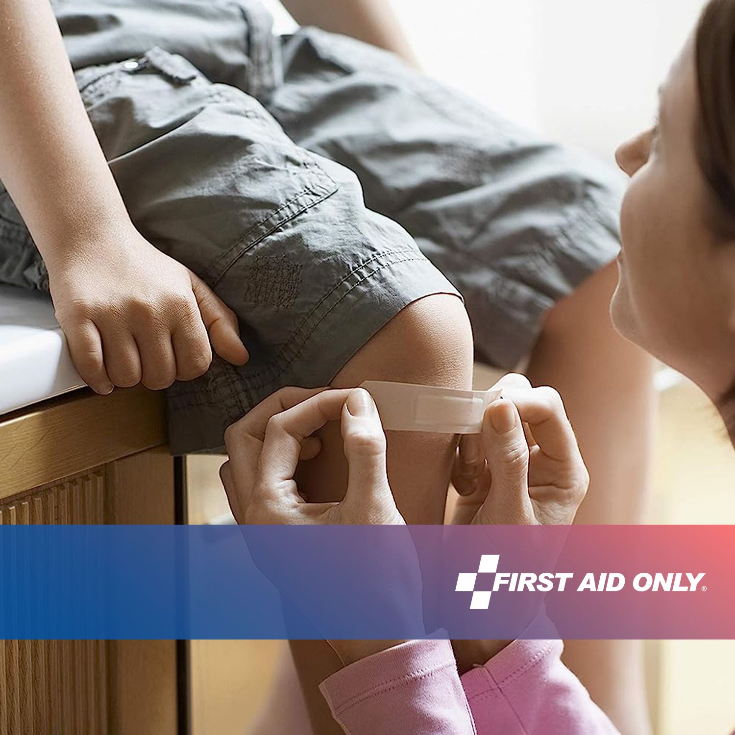 Kind mit Pflaster am Knie. Person legt Pflaster an. Marke: FIRST AID ONLY.